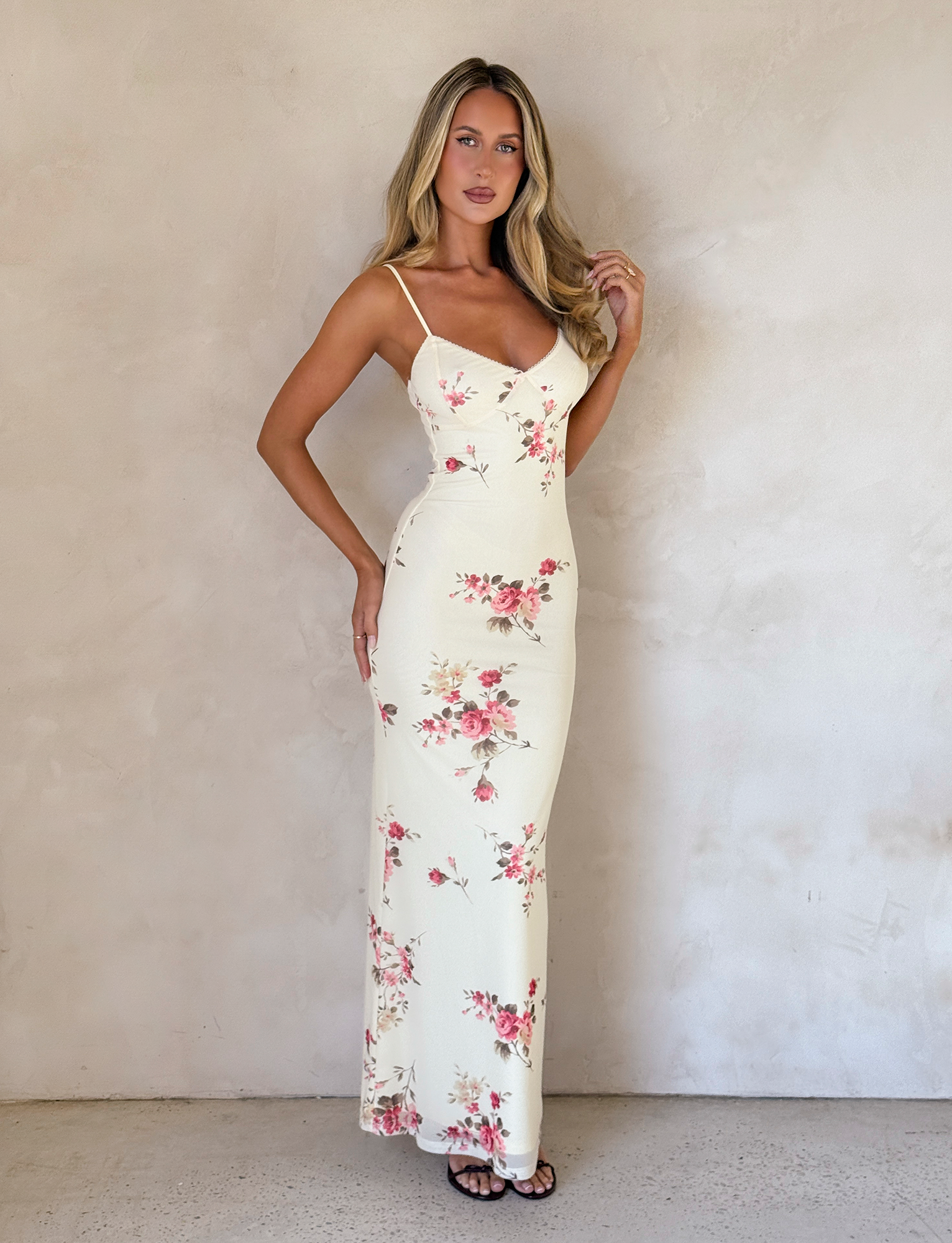A woman with long blonde hair poses by a beige wall, wearing the TIGERMIST JULIANNA DRESS in yellow macro pop floral print and black sandals, with one hand near her face.