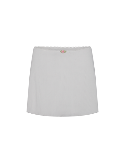 The MILANA SWIM SKIRT by TIGERMIST is a white ribbed mini skirt featuring a scalloped waistband and a small pink and green embroidered rose at the center, crafted from rib swim fabrication. Displayed on a white background.