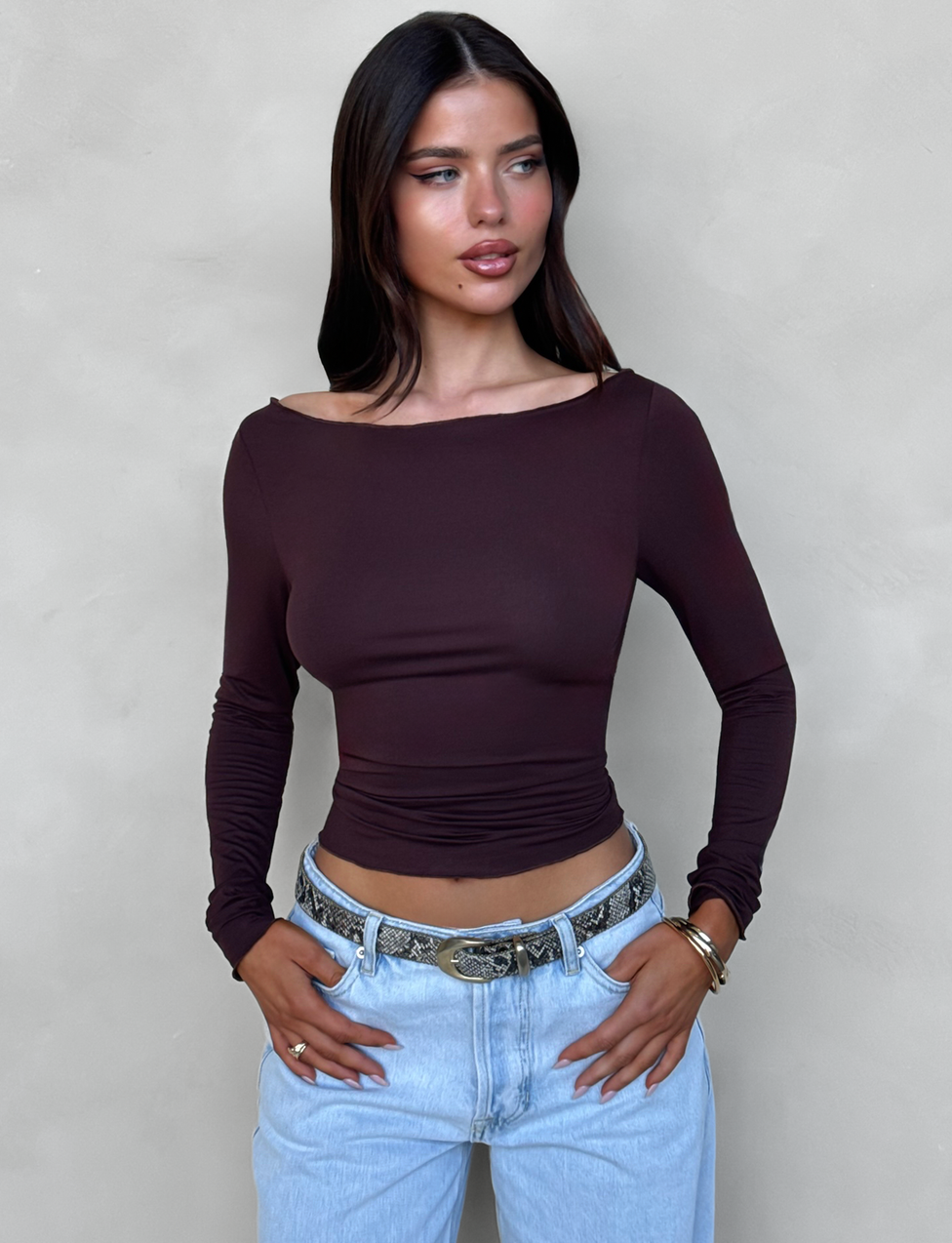 A woman with long, dark hair poses against a light grey textured background. She is wearing a fitted, off-the-shoulder TIGERMIST BIANKA TOP in a rich chocolate brown colorway, paired with light blue jeans featuring a detailed belt. With her hands in her pockets, she gazes to her left, embodying a chic bodycon silhouette.