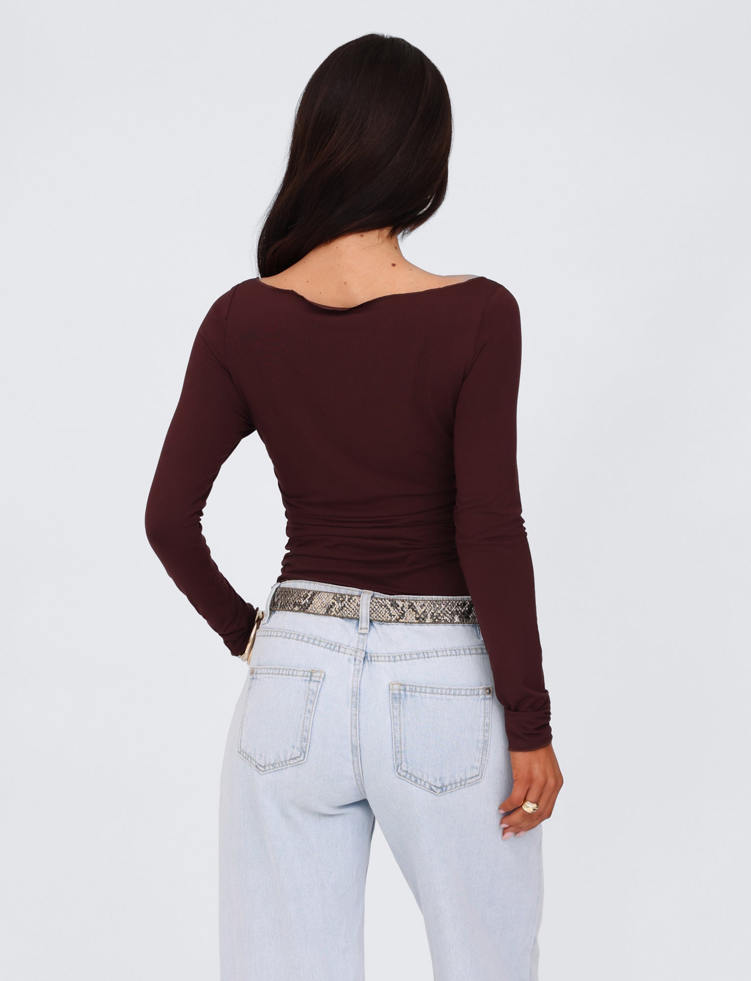 A person with long dark hair stands facing away from the camera. They are wearing a long-sleeved, chocolate brown TIGERMIST Bianka top and light blue jeans with a snakeskin-patterned belt. The background is plain and light.