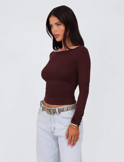 A woman with long dark hair stands against a plain background, wearing a chocolate brown Bianka top from Tigermist that features a bodycon silhouette. She pairs it with light blue high-waisted jeans and accessorizes with a snake-print belt and several bracelets on her wrist. She looks to her left with a serious expression.