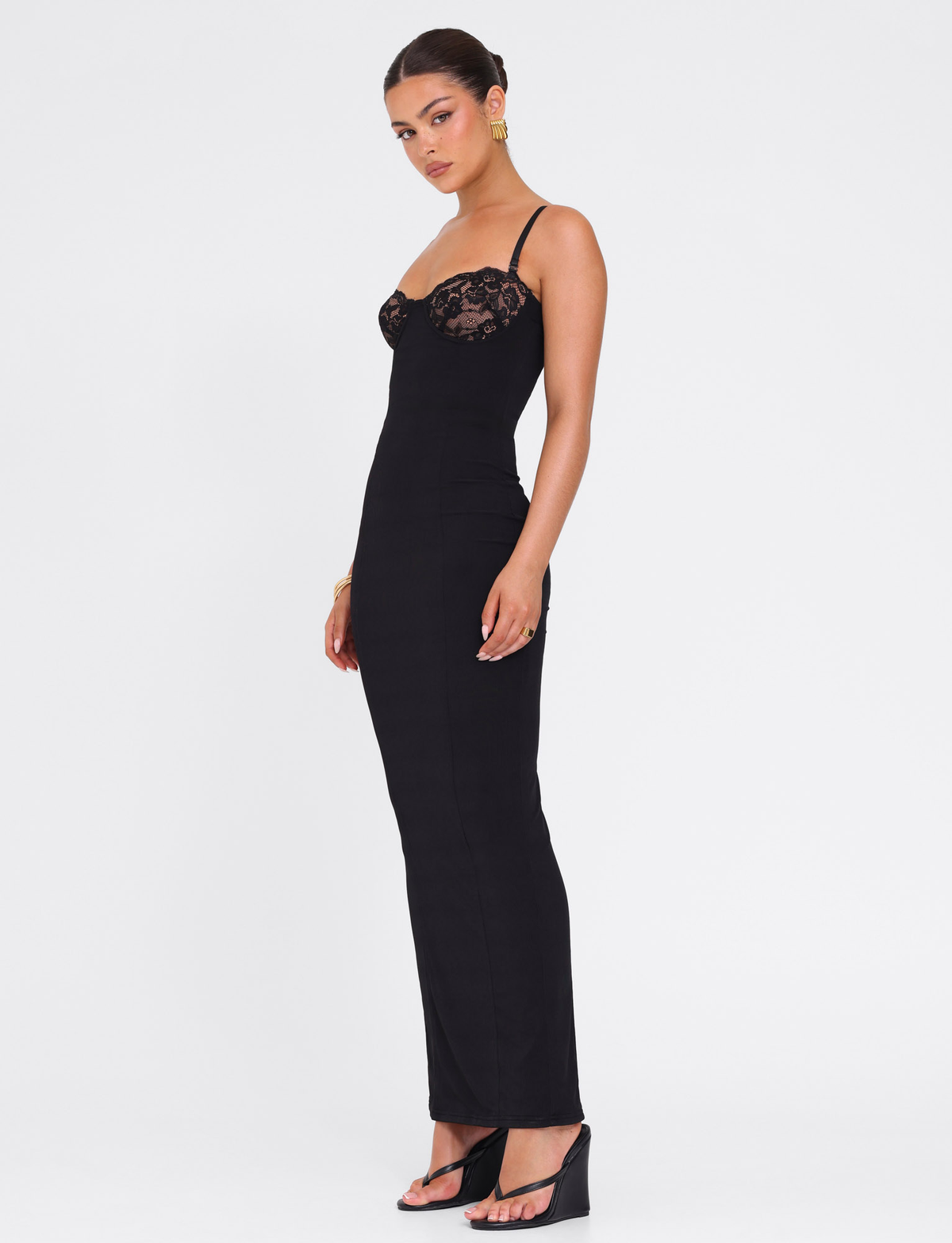 A woman stands sideways wearing the TIGERMIST ALLURA MAXI DRESS in black with lace bra cups, spaghetti straps, and black open-toe heeled sandals. Her hair is up, accessorized with gold earrings and a bracelet.
