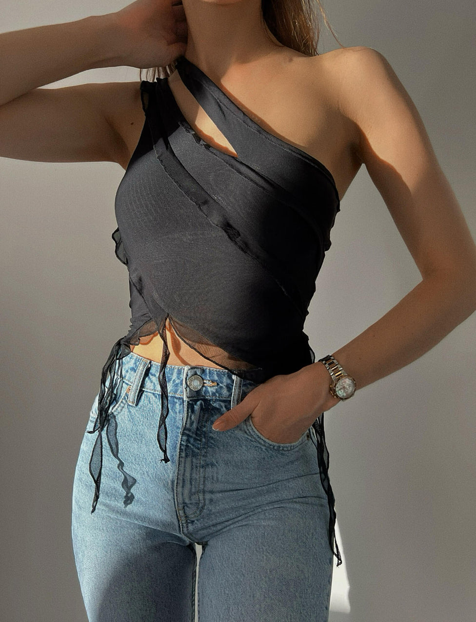 A person is wearing the stylish DIONNE TOP - BLACK from TIGERMIST, featuring asymmetrical crisscrossing straps and distressed edges, paired with high-waisted blue jeans. They have one hand in their pocket and the other resting near their head, showcasing a silver watch on their wrist.