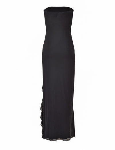 The Theo Maxi Dress - Black by TigerMist is a sleek, strapless evening gown with a floor-length design. It features a straight neckline and subtle ruffles cascading down one side, adding an elegant touch. The fabric is smooth and fitted, accentuating a sophisticated silhouette.