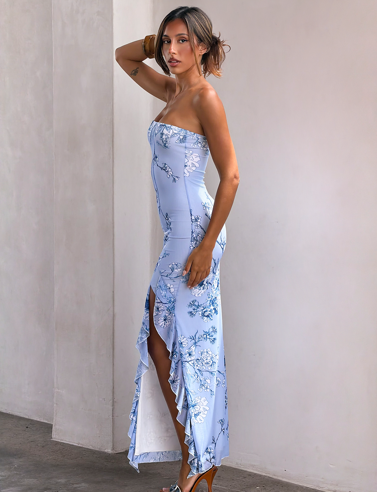 A woman models the TIGERMIST THEO MAXI DRESS - BLUE FLORAL, featuring mesh fabric, a bandeau neckline, thigh-high split, and ruffled hem. She pairs it with high-heeled sandals and poses against a plain light wall.