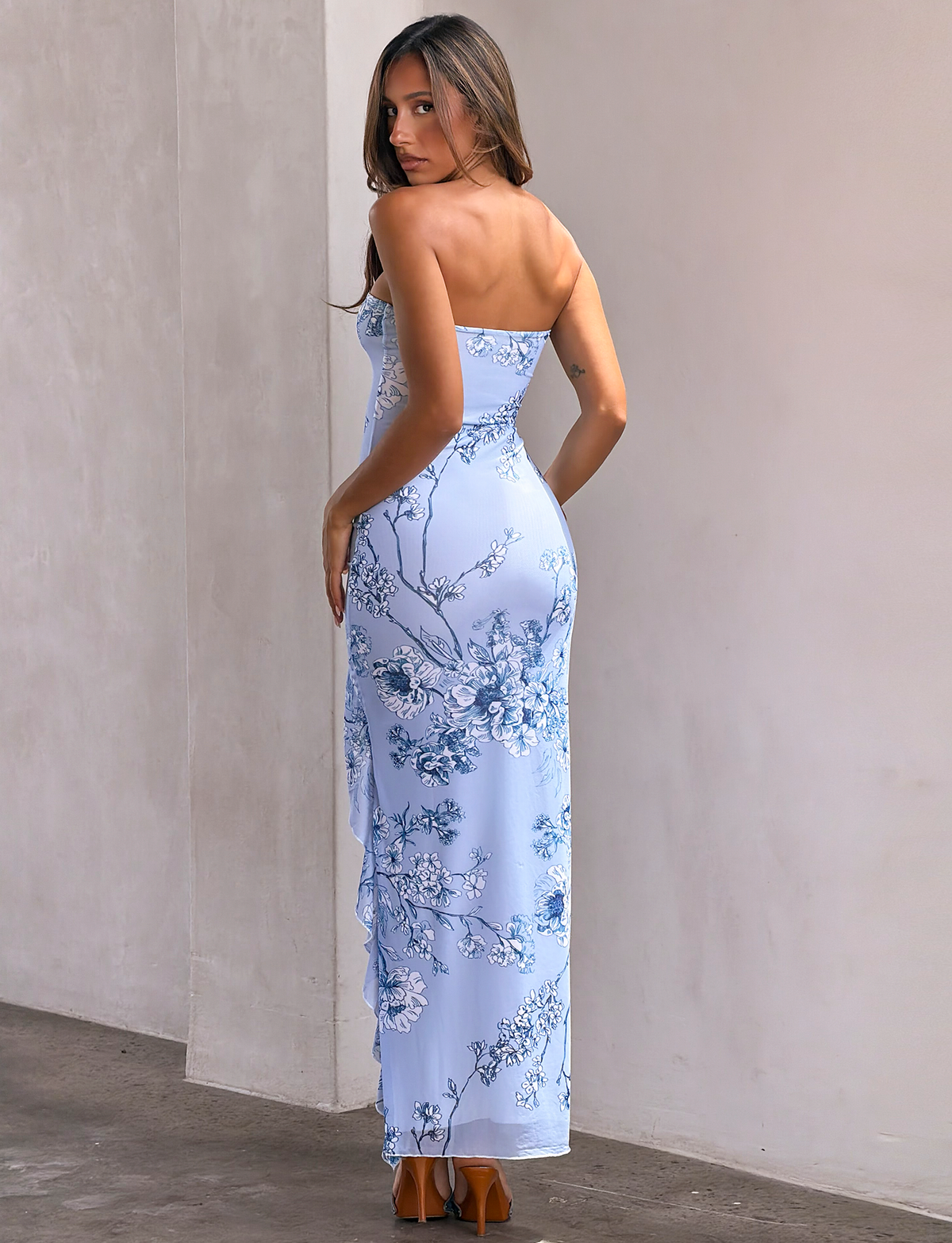 A woman in the TIGERMIST THEO MAXI DRESS - BLUE FLORAL, featuring mesh fabric, floral print, a bandeau top, and thigh-high split, pairs it with brown heels indoors. She faces away but glances over her shoulder at the camera. Light walls form the backdrop.