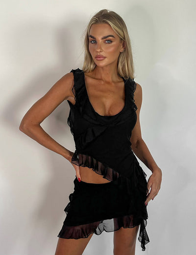 A woman with long blonde hair is posing against a plain white background. She is wearing the STORMI TOP - BLACK from TIGERMIST, featuring ruffled layers. Her right hand rests on her hip, and she gazes confidently forward.