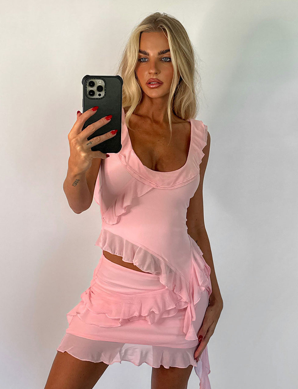 A person with long blonde hair takes a mirror selfie while wearing the STORMI TOP - PINK : BABY PINK from TIGERMIST. The light pink, ruffled, sleeveless top and matching skirt feature asymmetrical frill details. They hold a black smartphone with a visible rear camera against a plain white background.