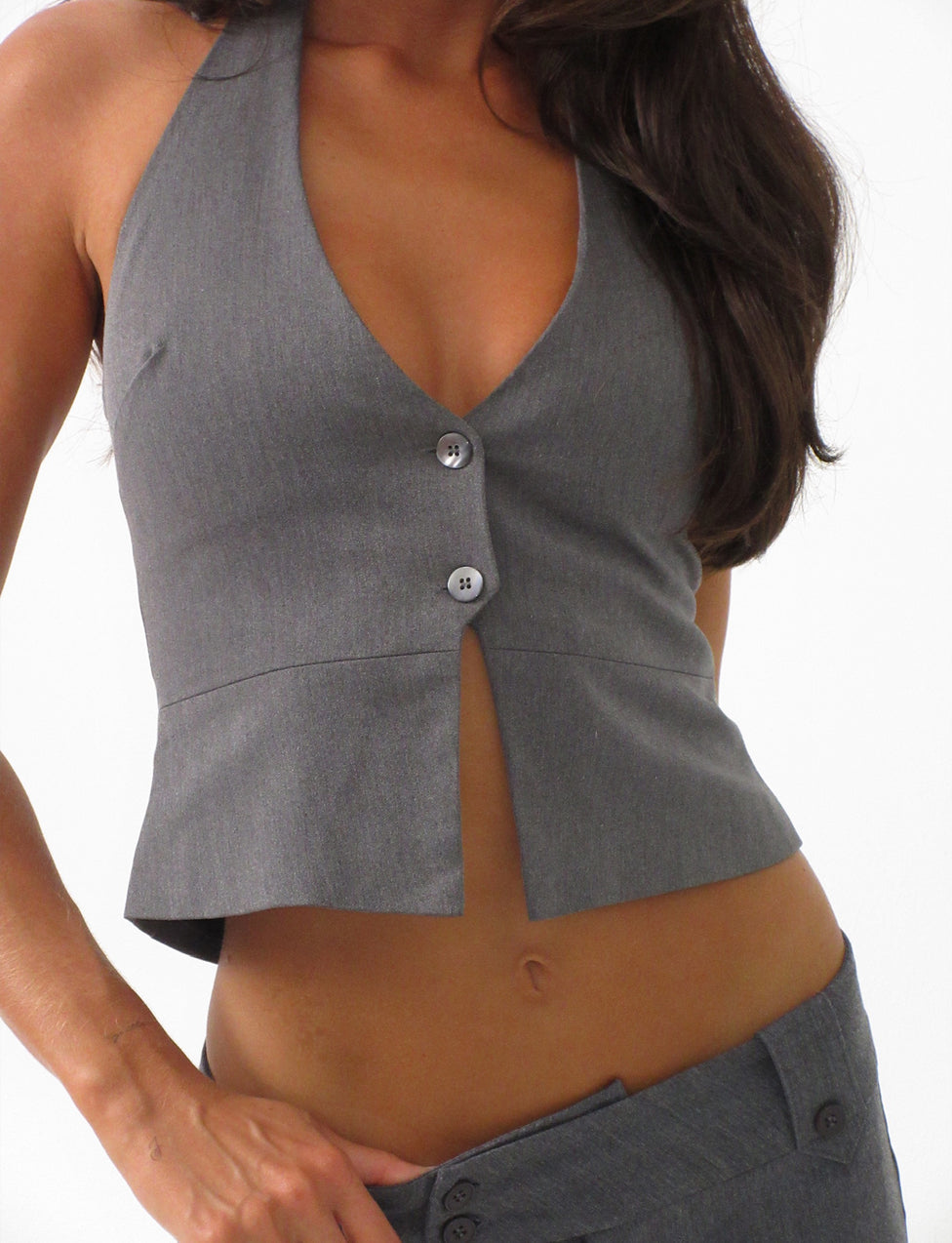 A person is wearing the TRISTAN VEST - GREY from TIGERMIST, which features a fitted design with two buttons down the front. The bottom button is left unfastened, creating a slight open slit. They are also wearing matching gray pants and have their hand resting on the waistband. Their long dark hair is draped over one shoulder.