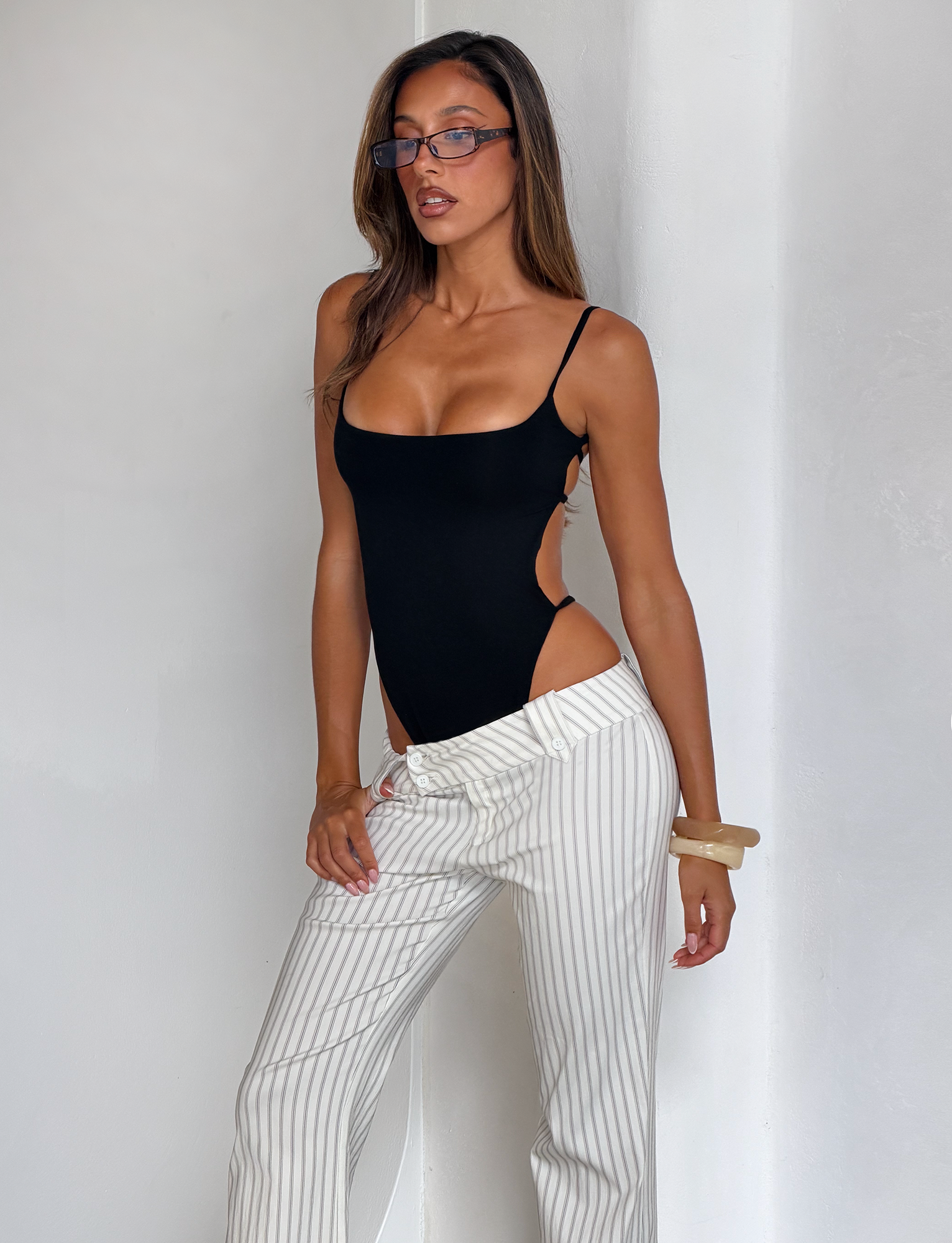 A woman wearing glasses and the TIGERMIST MINERVA BODYSUIT in black pairs it with white pinstripe pants, posing against a plain white wall with one hand in her pocket and the other resting on her leg.
