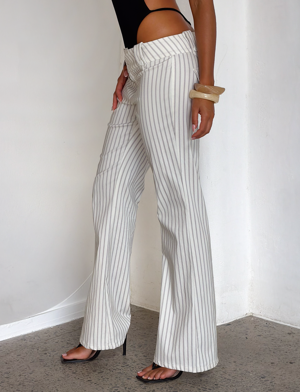 A woman stands against a white wall wearing the TIGERMIST KITTIE PANT in ivory stripe—a high-waisted, flared pant with thin gray stripes—paired with a black sleeveless top, black heels, and a chunky bangle.