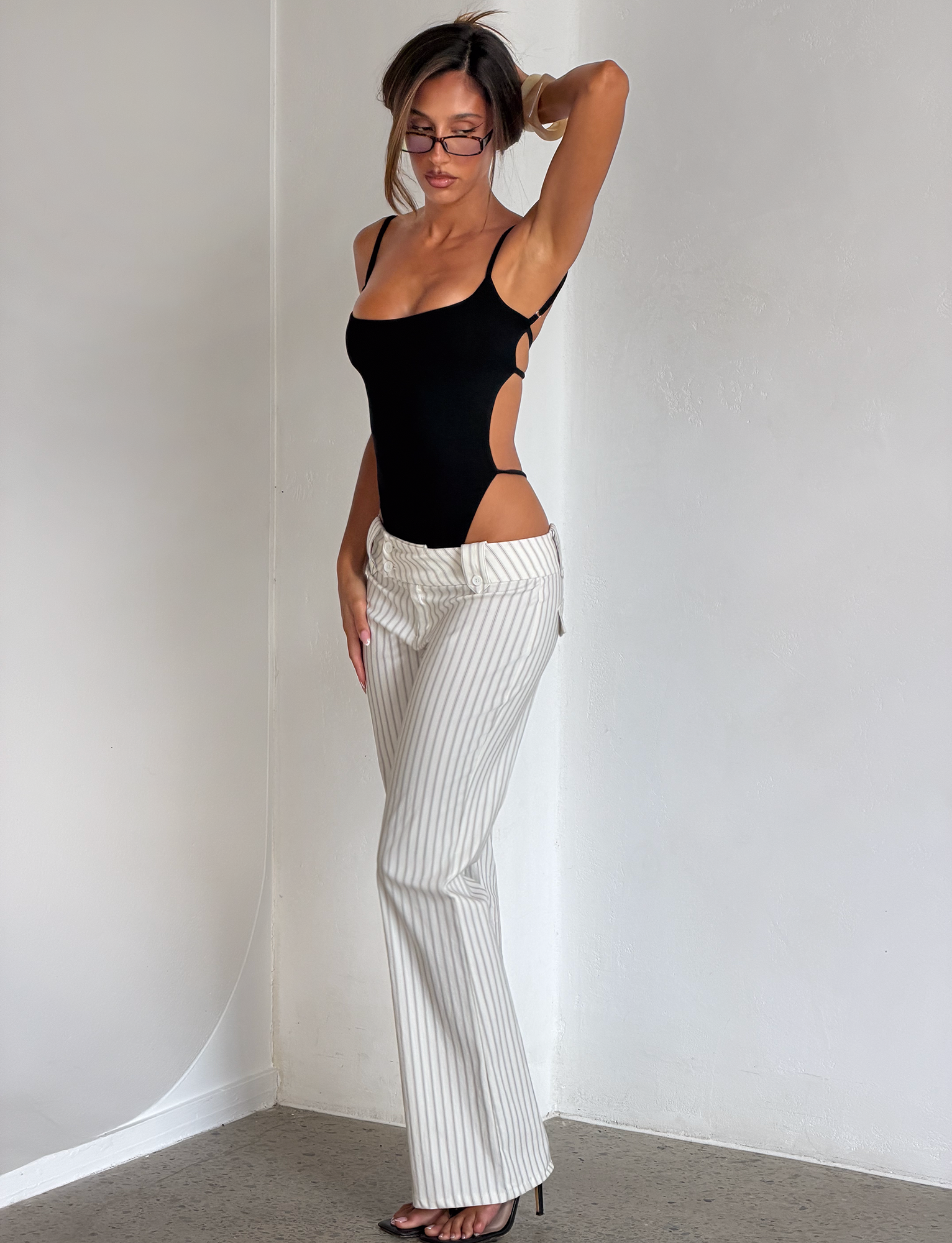 A woman poses indoors in the TIGERMIST MINERVA BODYSUIT - BLACK, paired with white pinstripe pants and black heels. She adjusts her glasses with one hand, the other arm raised, standing against a plain white wall.