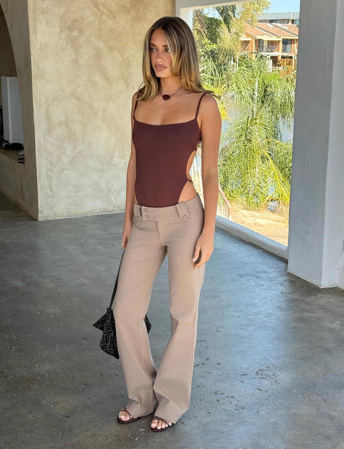 Indoors on a concrete floor, a woman wears the TIGERMIST KITTIE PANT in taupe paired with a brown sleeveless bodysuit and sandals. She holds a dark handbag, sunlight catches her wavy hair, and greenery is visible through the window behind her.