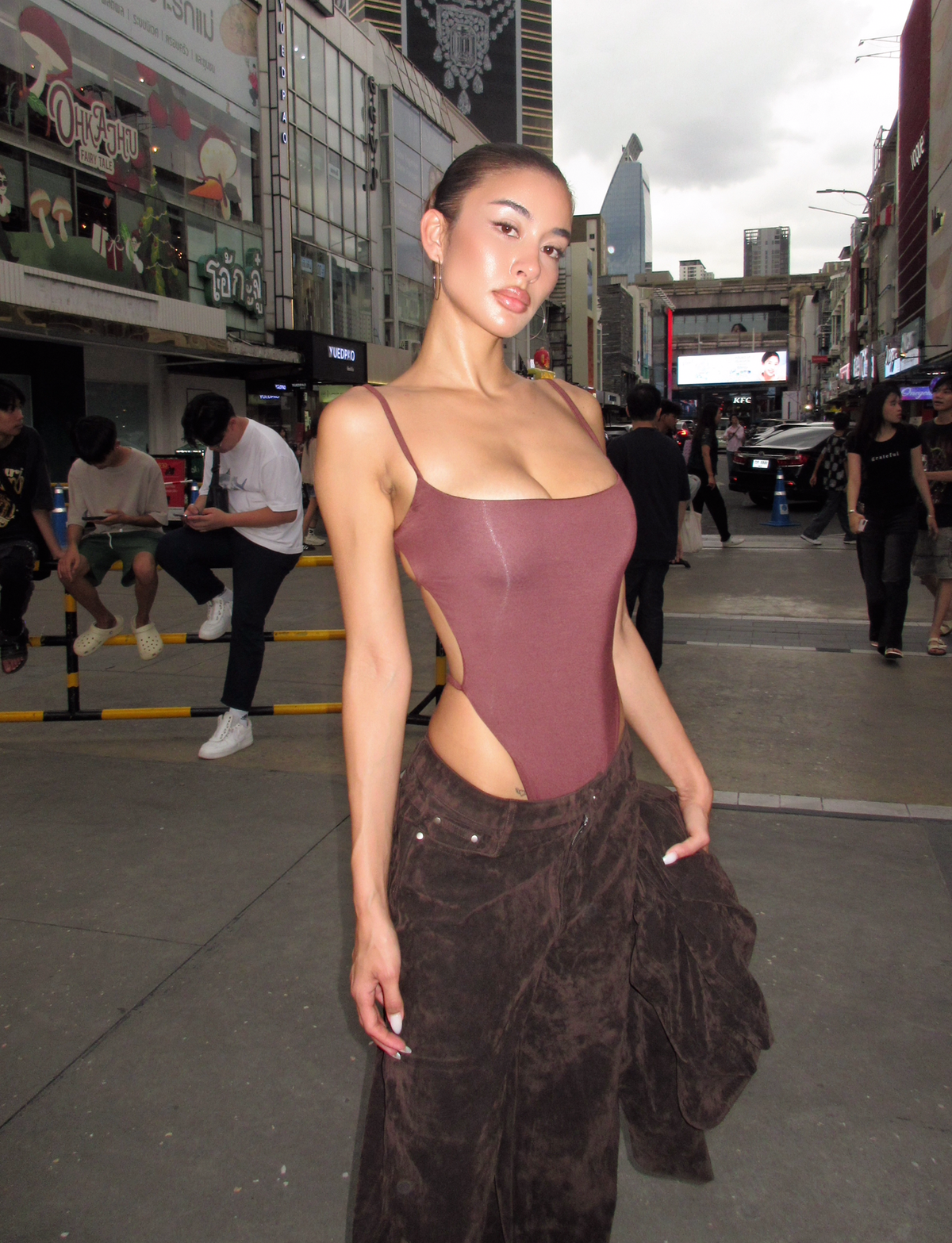 A woman wears the TIGERMIST MINERVA BODYSUIT in Chocolate, featuring adjustable shoulder straps and an open back, paired with loose brown pants as she poses confidently on a bustling city street under a cloudy sky.