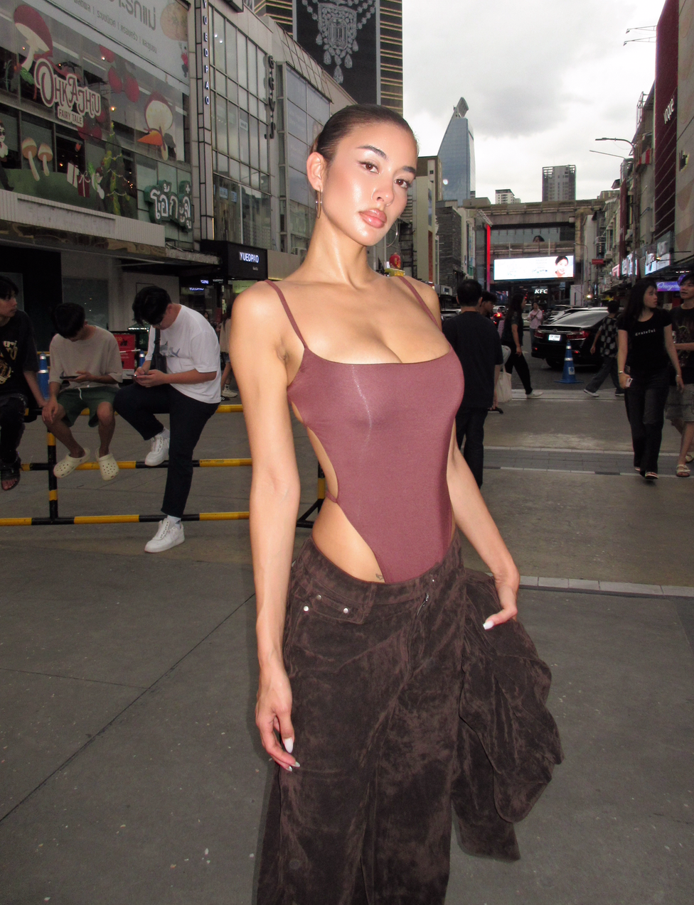 A woman wears the TIGERMIST MINERVA BODYSUIT in Chocolate, featuring adjustable shoulder straps and an open back, paired with loose brown pants as she poses confidently on a bustling city street under a cloudy sky.