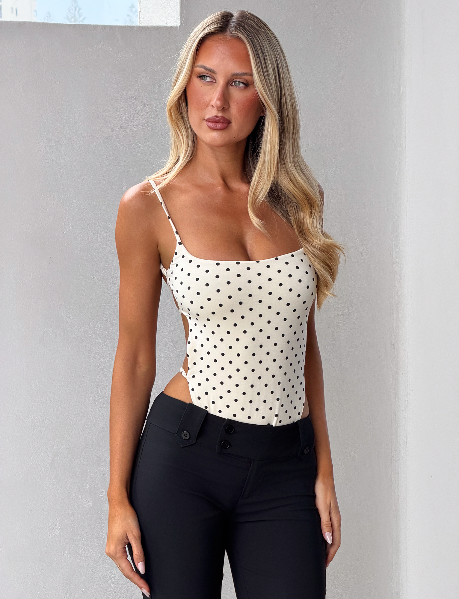 A woman with long blonde hair models the Tiger Mist CL - MINERVA BODYSUIT, a white polka dot sleeveless top, paired with black pants and stands against a plain light wall, gazing thoughtfully to the side.
