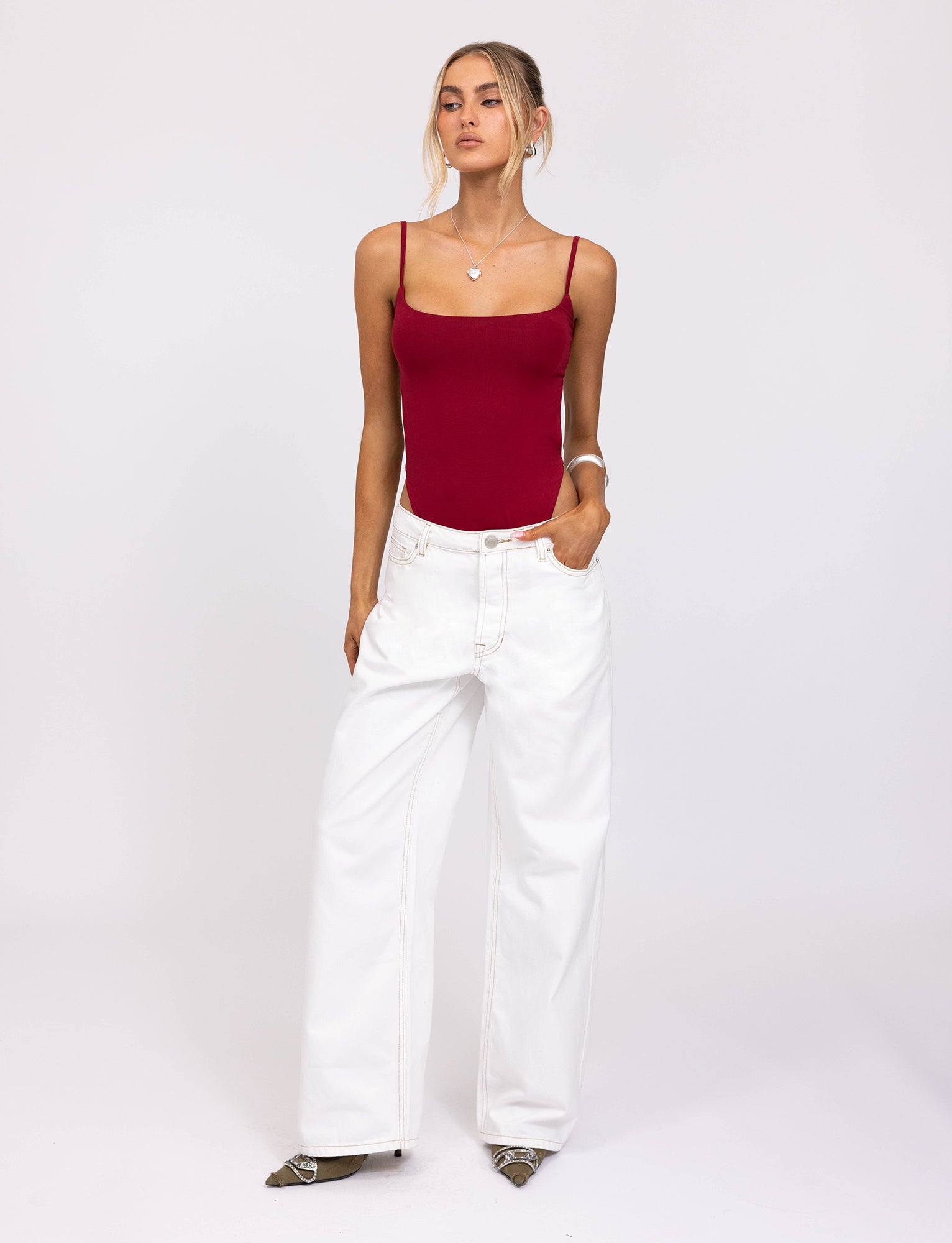 A person stands against a plain white background, dressed in a red MINERVA BODYSUIT by TIGERMIST and loose-fitting white jeans. Their hair is tied back in a loose bun, with one hand resting casually in their pocket. The outfit is completed with pointed shoes.