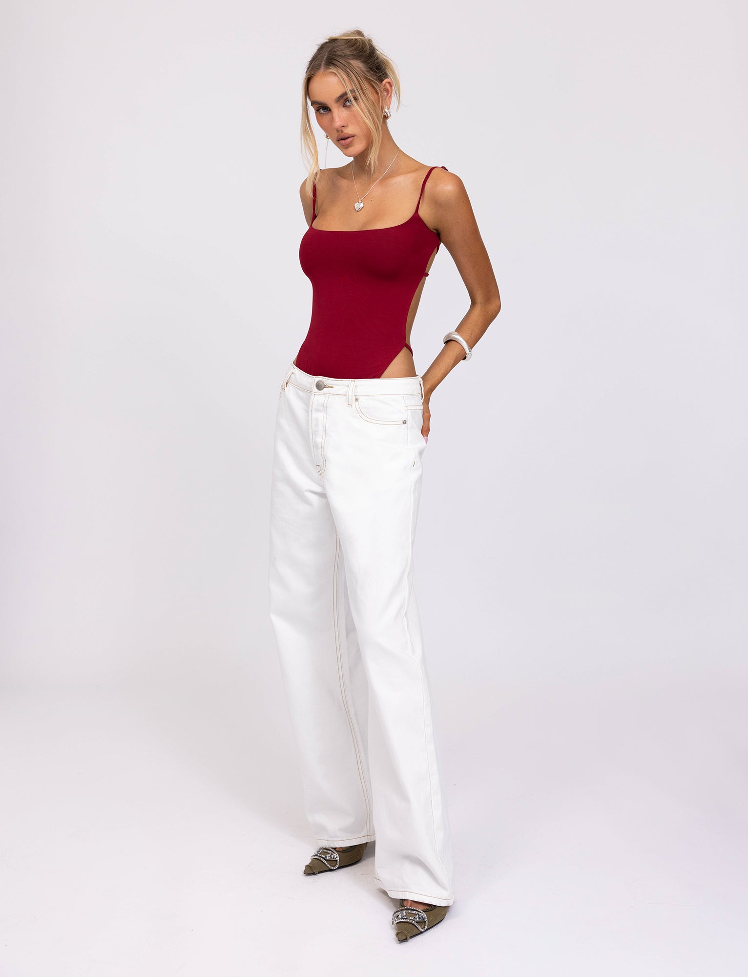 A woman with blonde hair stands against a plain white background. She wears the Minerva Bodysuit in red from TigerMist and high-waisted, wide-legged white jeans. She accessorizes with jewelry and gray pointed shoes. Her posture is relaxed, with one hand in her pocket.