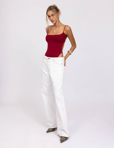 A woman with blonde hair stands against a plain white background. She wears the Minerva Bodysuit in red from TigerMist and high-waisted, wide-legged white jeans. She accessorizes with jewelry and gray pointed shoes. Her posture is relaxed, with one hand in her pocket.