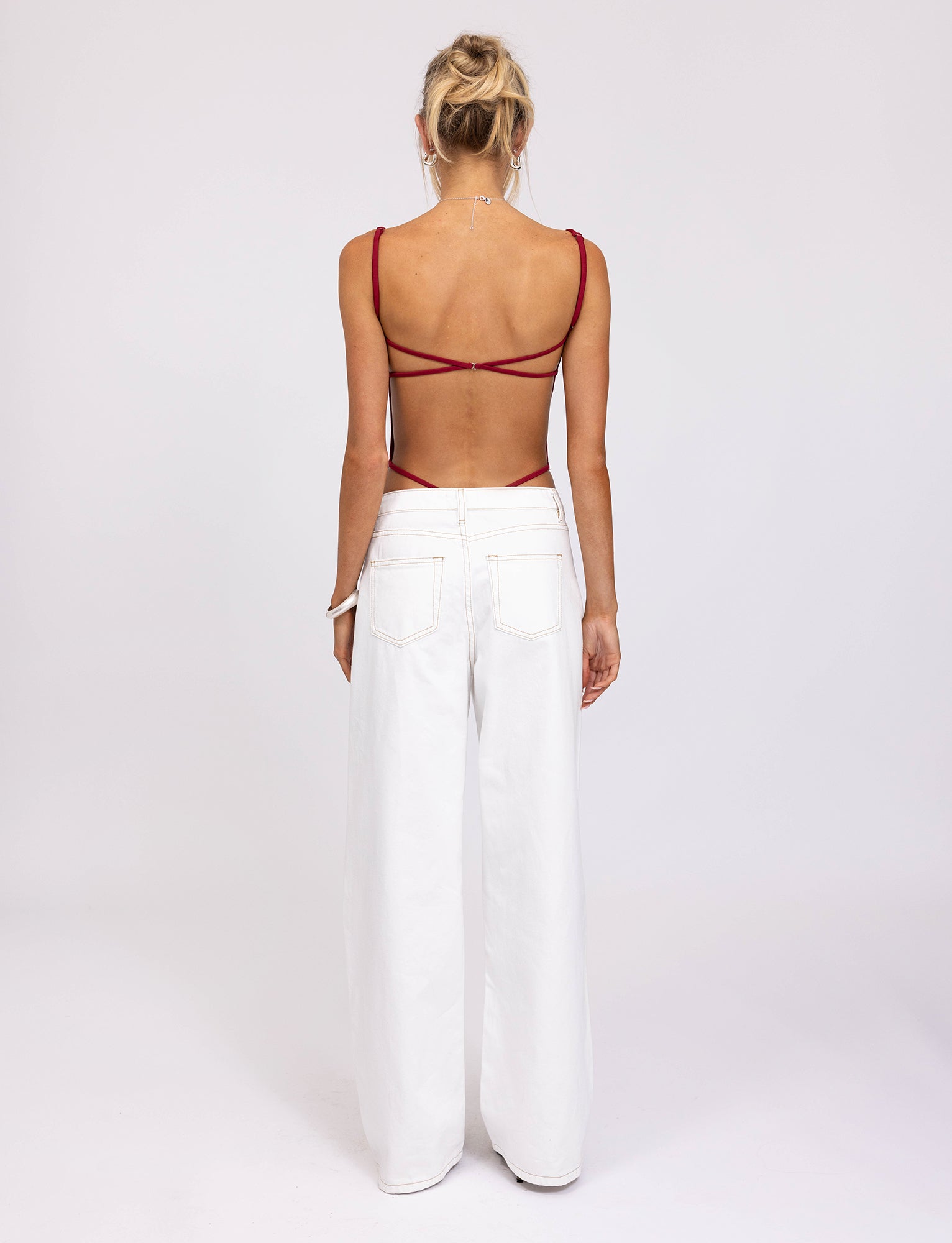 A person with blonde hair is standing with their back to the camera, showcasing a chic TIGERMIST MINERVA BODYSUIT in red, paired with white, wide-leg pants. The plain white background enhances the stylish ensemble.