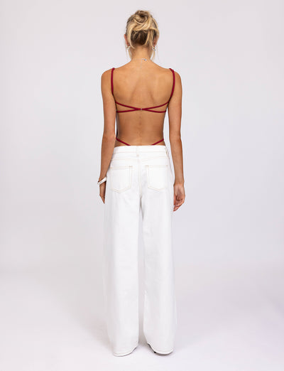 A person with blonde hair is standing with their back to the camera, showcasing a chic TIGERMIST MINERVA BODYSUIT in red, paired with white, wide-leg pants. The plain white background enhances the stylish ensemble.