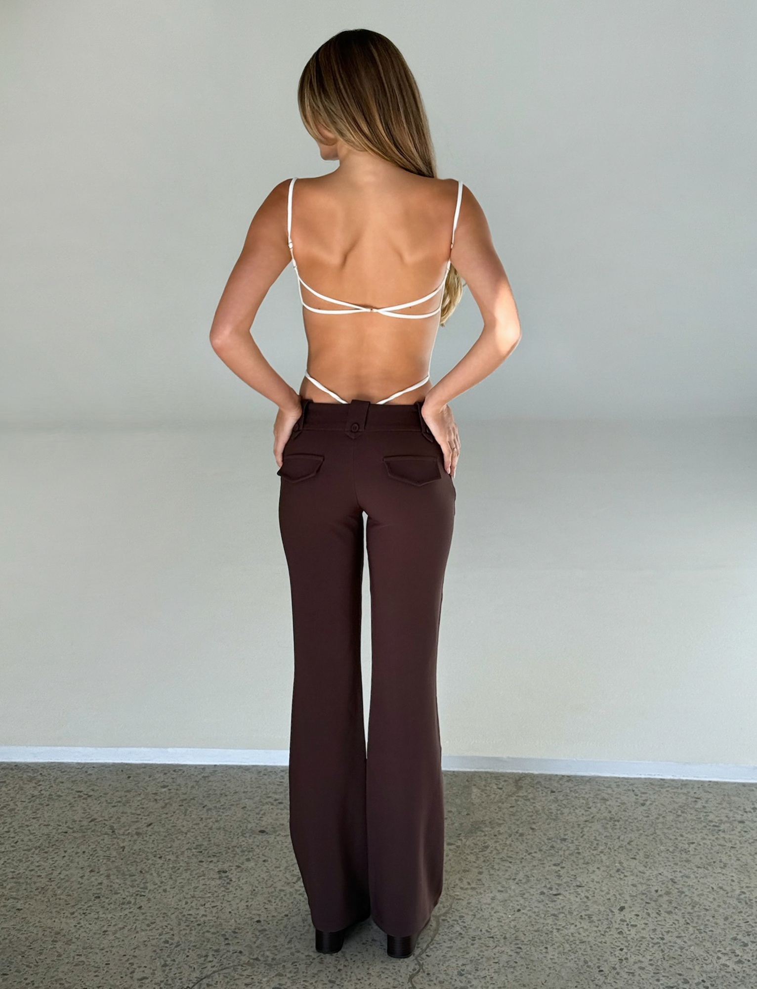 A woman stands with her back to the camera in a strappy white top and TIGERMIST KITTIE PANT - BROWN : CHOCOLATE, featuring a high-waisted, flared fit. Her hands rest on her hips as her long hair flows down her back.