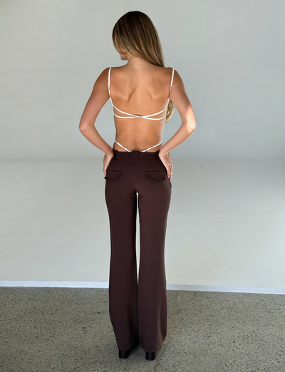 A woman stands with her back to the camera in a strappy white top and TIGERMIST KITTIE PANT - BROWN : CHOCOLATE, featuring a high-waisted, flared fit. Her hands rest on her hips as her long hair flows down her back.