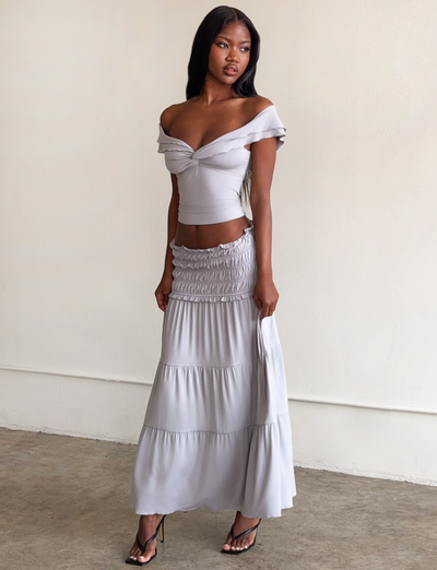 A woman with long dark hair stands indoors against a light wall, wearing an off-the-shoulder fitted crop top and the TIGERMIST DENALI MAXI SKIRT in Dove Grey, both in soft modal fabric with ruched details, paired with black high-heeled sandals.