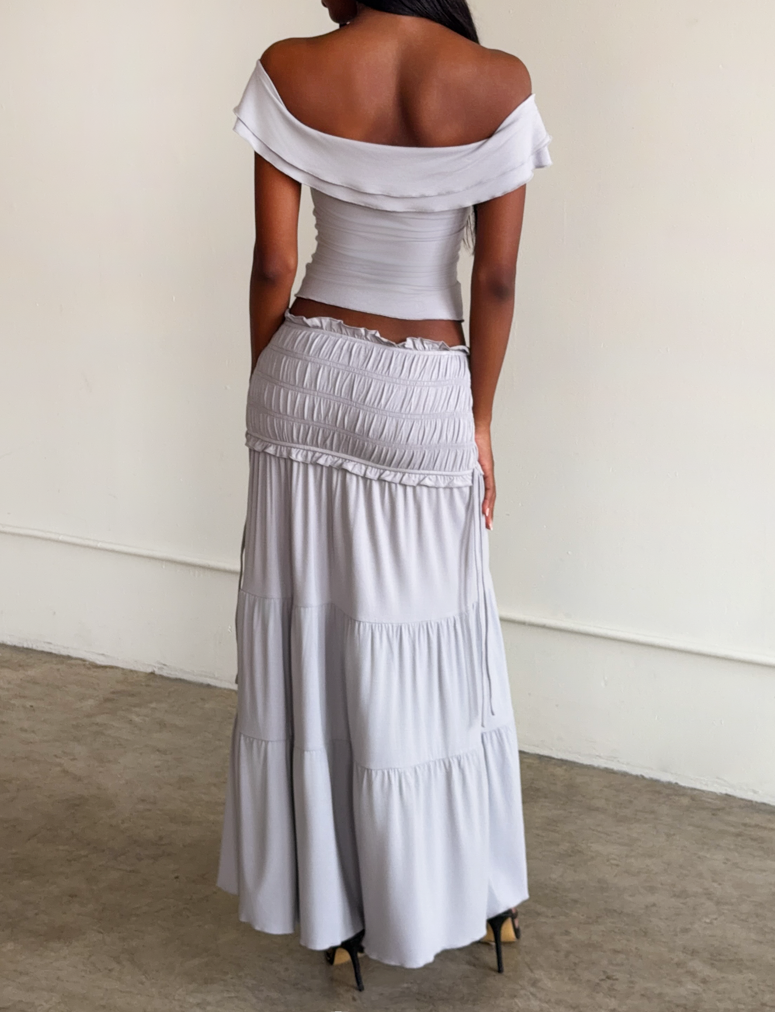 A woman stands indoors facing away, wearing an off-the-shoulder fitted top and the TIGERMIST DENALI MAXI SKIRT - DOVE GREY, a flowy modal maxi skirt. The background is plain and minimalistic.