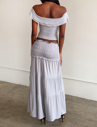 A woman stands indoors facing away, wearing an off-the-shoulder fitted top and the TIGERMIST DENALI MAXI SKIRT - DOVE GREY, a flowy modal maxi skirt. The background is plain and minimalistic.