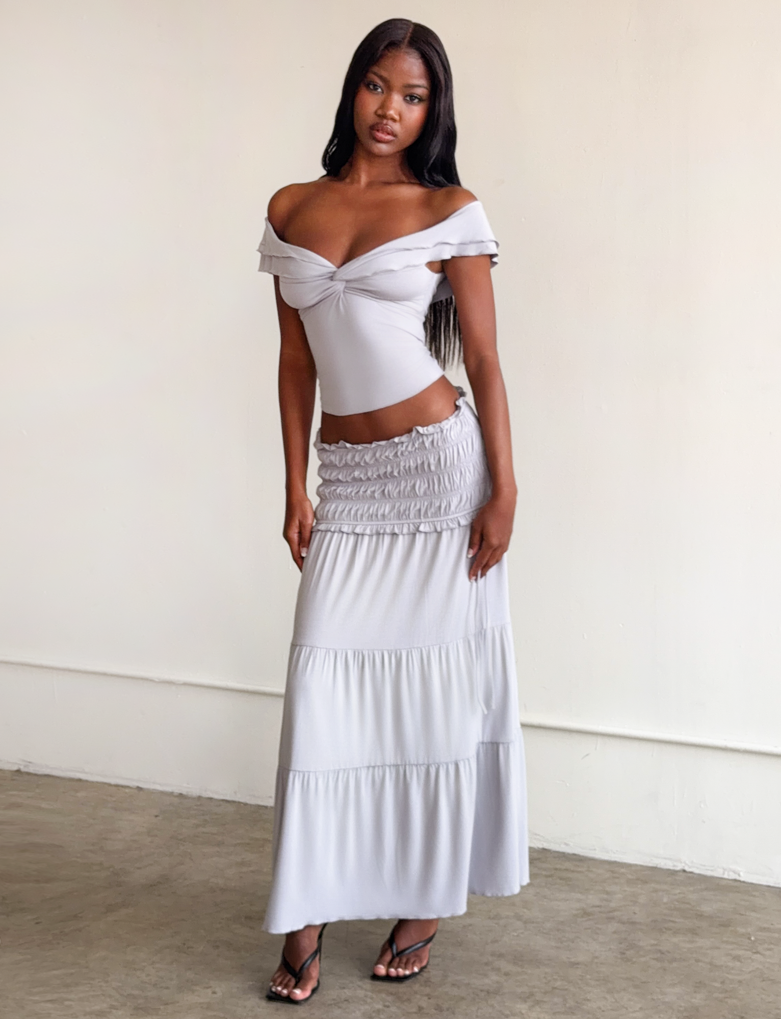 A woman with long dark hair wears the TIGERMIST DENALI TOP in Dove Grey, an off-the-shoulder modal top, and a matching tiered skirt. She stands indoors against a plain wall, styled with black heeled sandals.