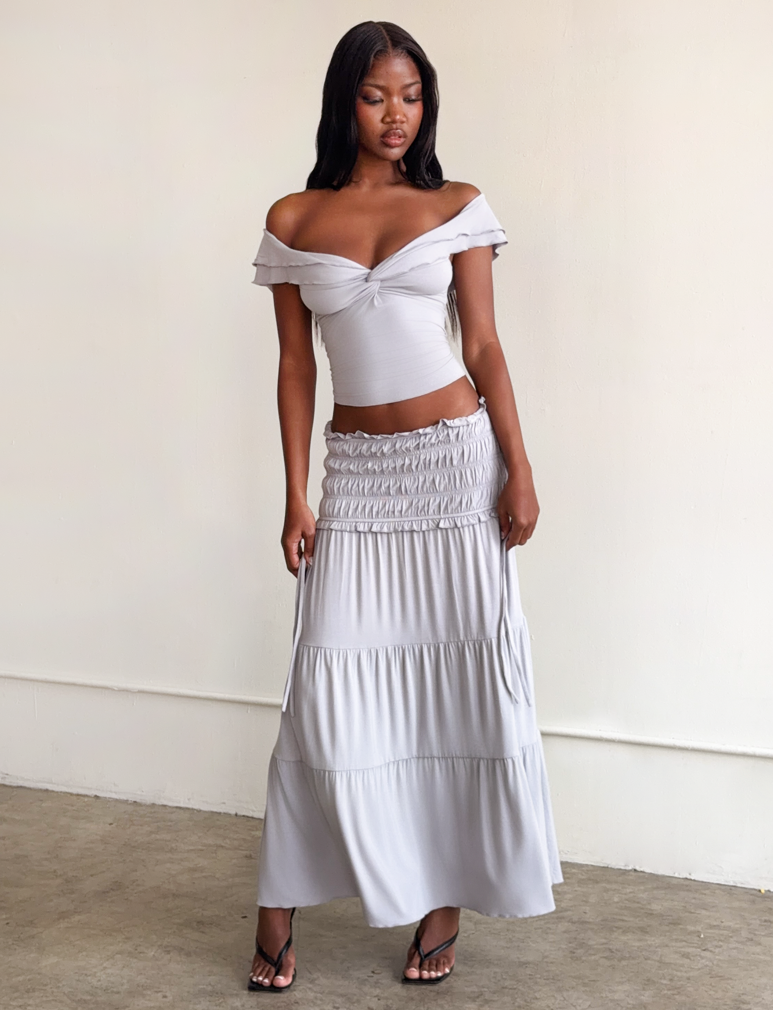 A woman stands indoors on a concrete floor, looking down with one hand by her side, wearing a light gray off-the-shoulder crop top and the TIGERMIST DENALI MAXI SKIRT in Dove Grey crafted from soft modal fabric.