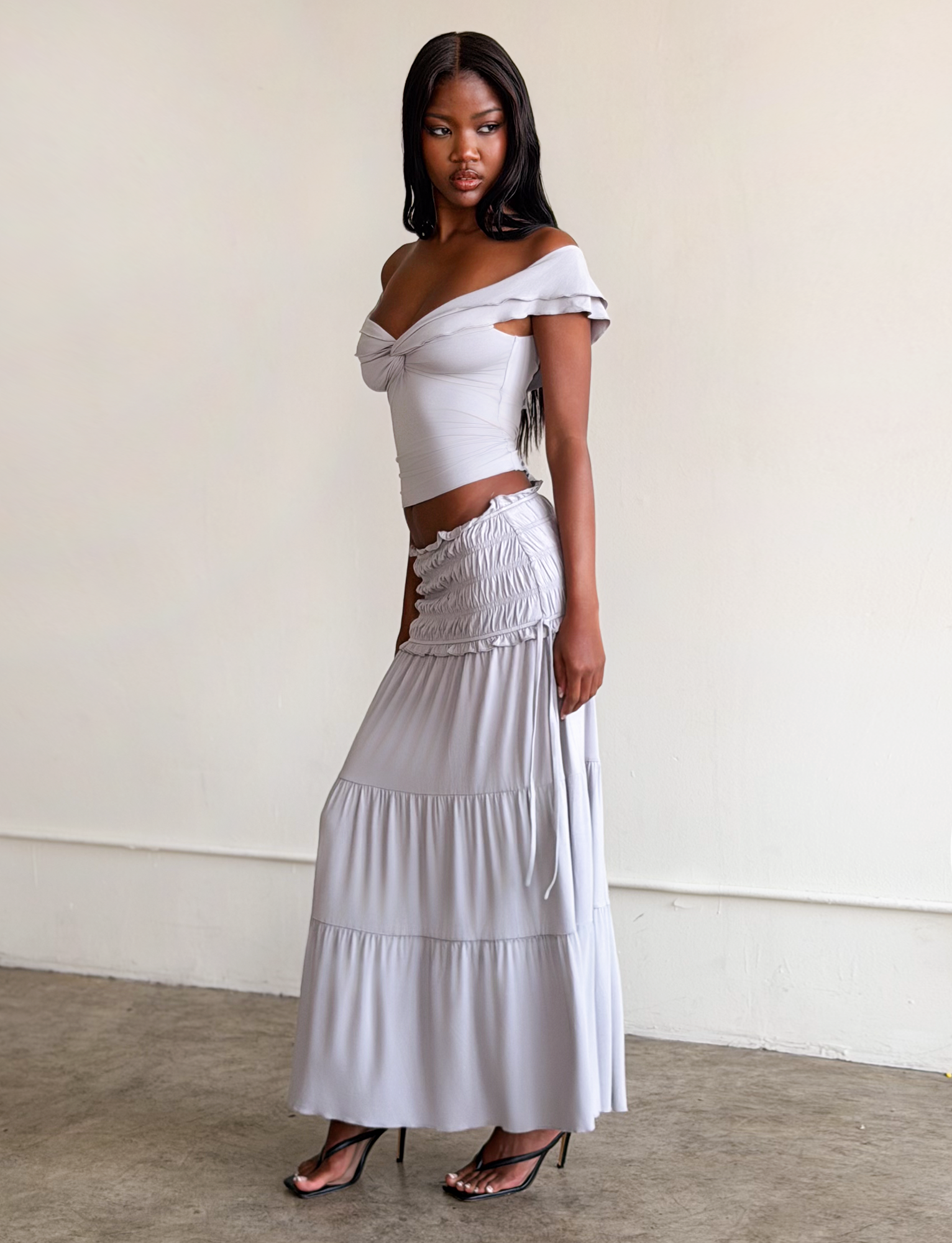 A woman poses indoors in a light gray off-shoulder top and the TIGERMIST DENALI MAXI SKIRT - DOVE GREY, featuring ruched details, styled with black heeled sandals against a plain background.