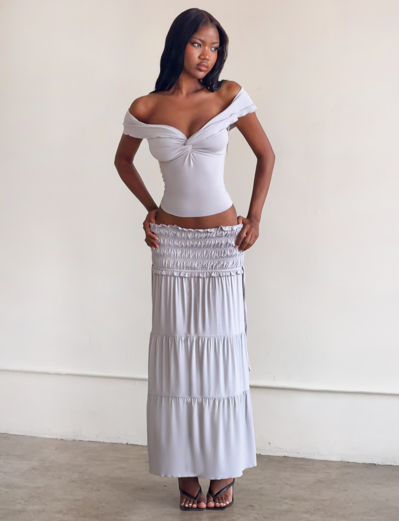 A woman stands indoors in a fitted white off-shoulder top and the TIGERMIST DENALI MAXI SKIRT in Dove Grey, posing confidently with one hand on her hip against a neutral background.