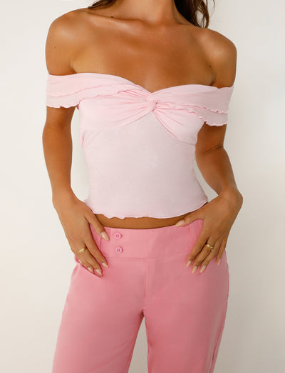 A person is wearing the DENALI TOP - PINK by TIGERMIST, an off-the-shoulder light pink top with a twist knot in the center. They have paired it with matching Tiger Mist pink pants that feature a three-button detail on the waistband. The person's right hand is resting on their hip while their left hand hangs by their side.