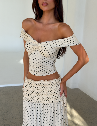 A woman poses indoors with one hand on her hip, wearing the TIGERMIST DENALI TOP - CREAM DOT, an off-shoulder cream top with black polka dots, paired with a matching ruffled high-waisted skirt. Her face is partially out of frame.