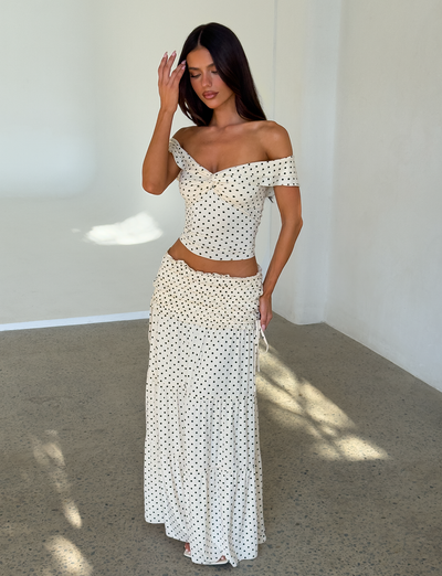 A woman stands indoors in the TIGERMIST DENALI TOP - CREAM DOT, an off-shoulder top with black polka dots, paired with a matching long skirt. She has long dark hair and touches her forehead with one hand.