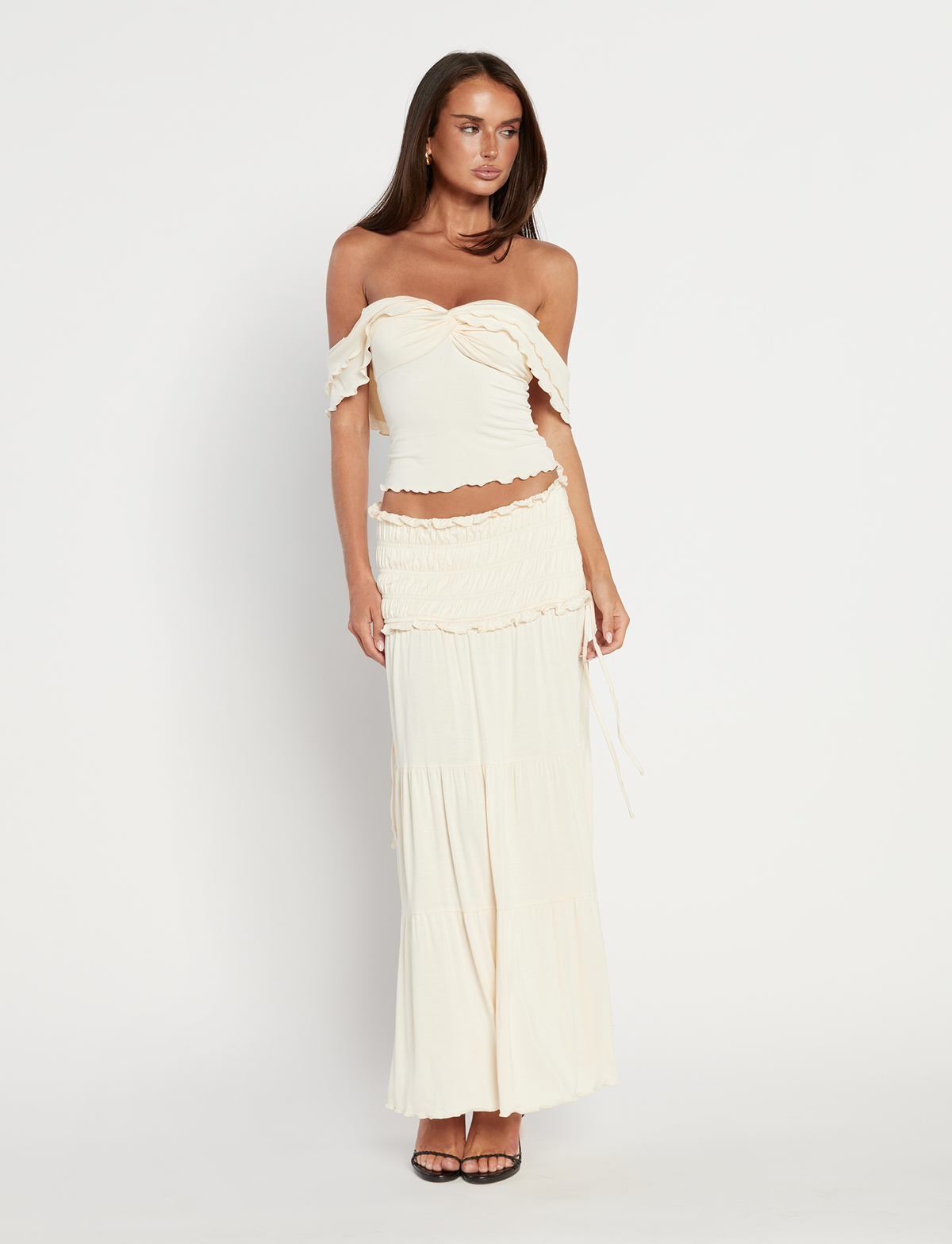 A woman with long brown hair wears the TIGERMIST DENALI MAXI SKIRT in Neutral: Bone, paired with an off-shoulder cream ruffled crop top, standing against a white background and styled with black heeled sandals.
