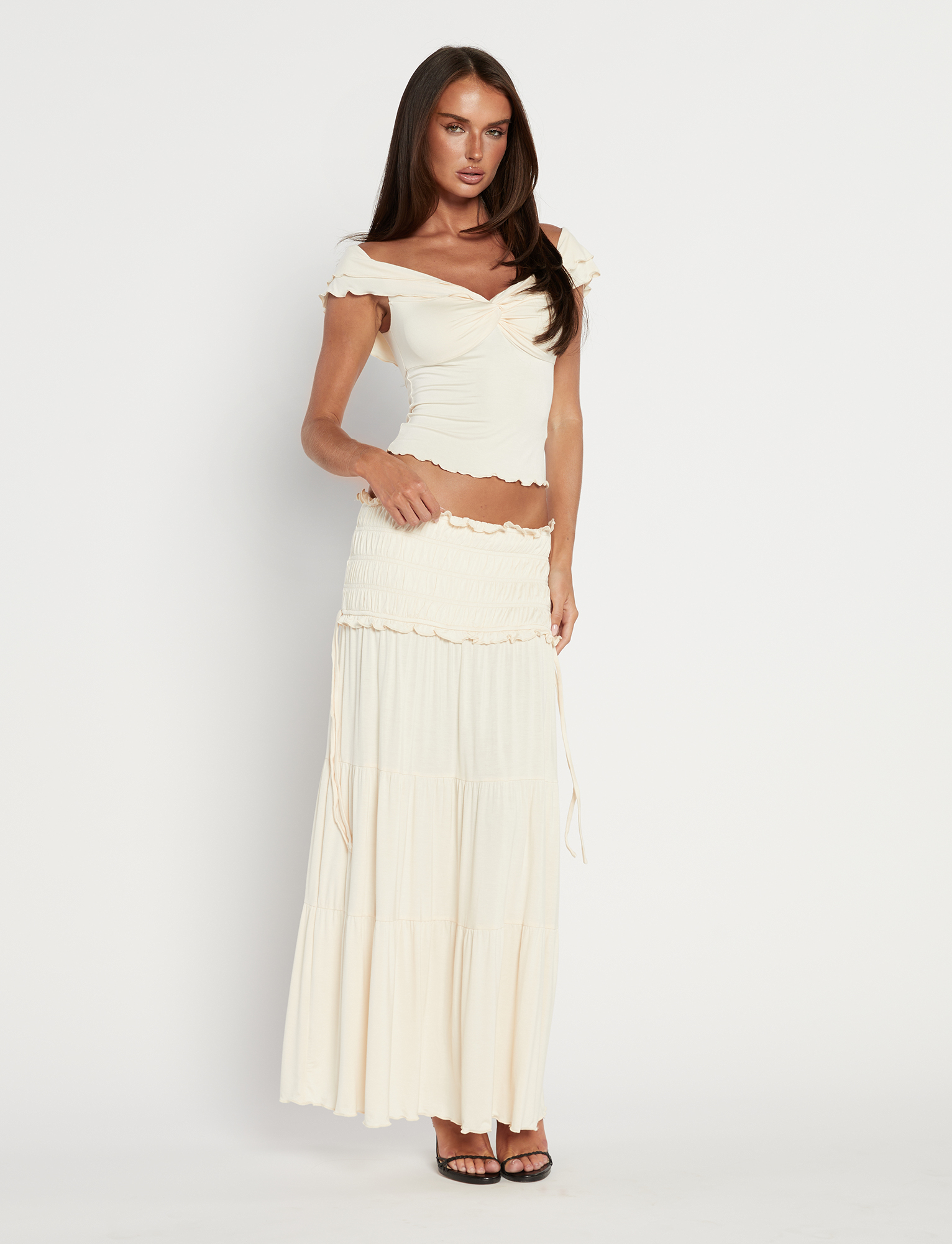 A woman with long brown hair models the TIGERMIST DENALI MAXI SKIRT in Bone, paired with a matching off-shoulder cream crop top, featuring a shirred waistband and soft buttery jersey, posing against a white background.