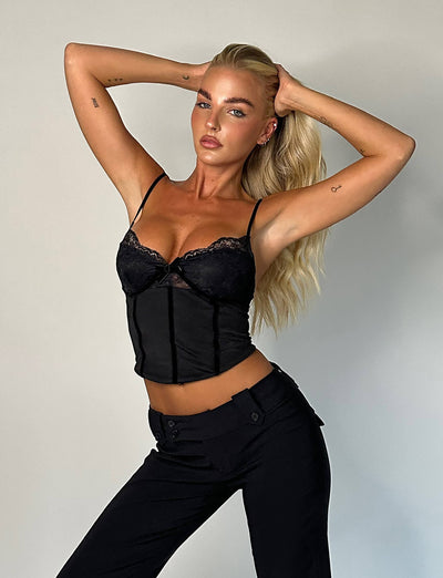 A person with long blonde hair poses against a plain background, clad in the MORENA TOP - BLACK and black pants from TIGERMIST. With hands raised and fingers running through their hair, they gaze confidently ahead, showcasing the tattoos on their arms.