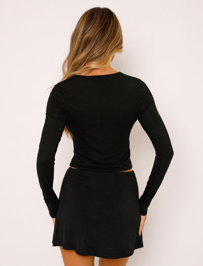 A woman with long, wavy hair faces away from the camera, wearing a black long-sleeve top and the TIGERMIST ALANI SKIRT in black. The background is plain and light-colored, giving the scene a minimalist Tiger Mist vibe.