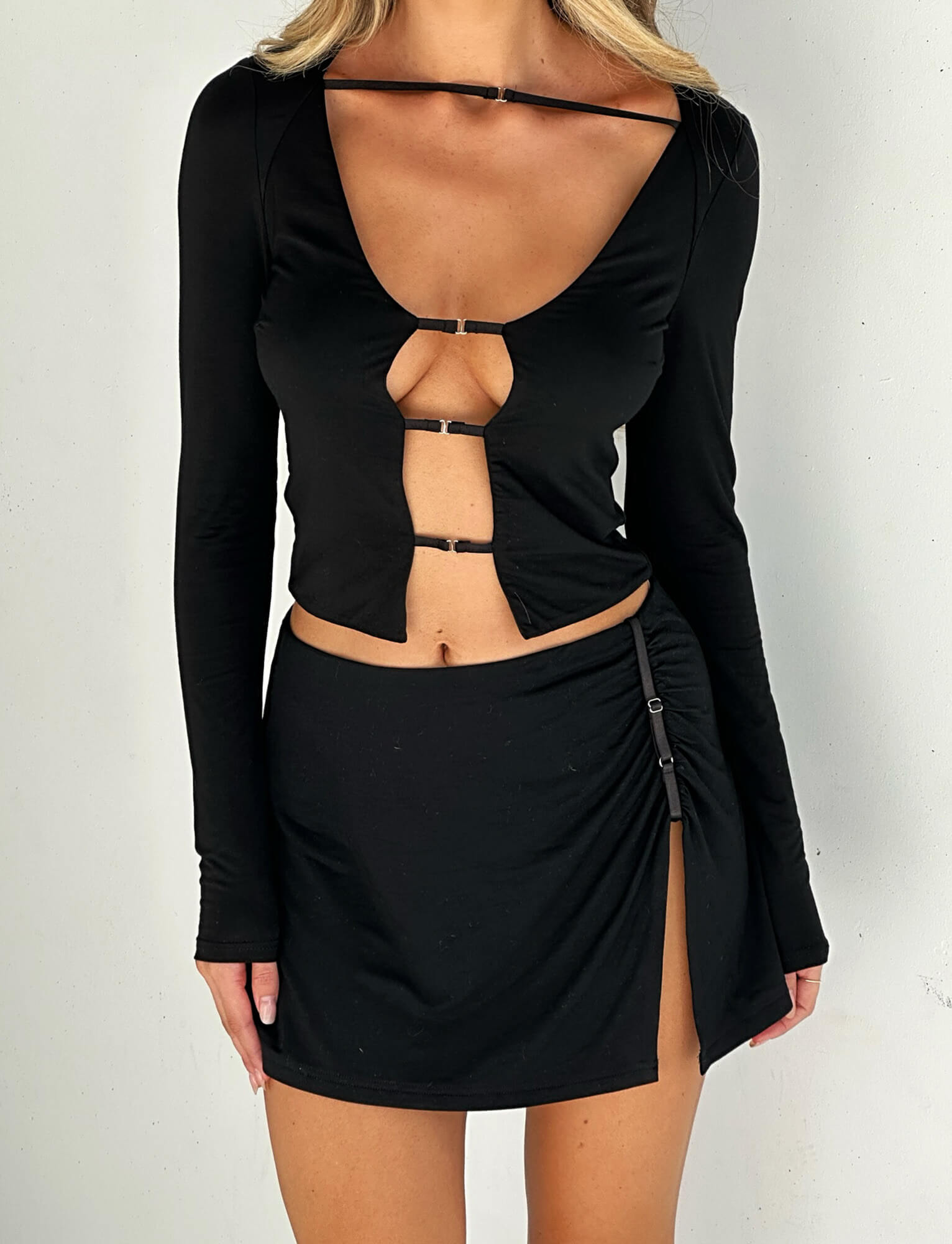 A person is wearing a chic ensemble from TIGERMIST. The black top boasts long sleeves with three front cut-out sections, linked by small metallic connectors, while the ALANI SKIRT - BLACK features a high slit on one side, secured by a small buckle.