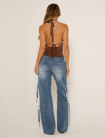 A woman stands with her back to the camera, wearing a stylish brown halter top from Tiger Mist and blue BAINES PANT - BLUE : LIGHT WASH wide-legged jeans with multiple pockets and zippers. Her long wavy hair cascades down her back against a solid, light-colored wall.