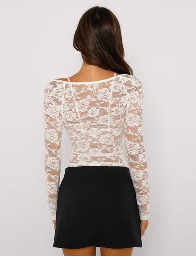A woman with long brown hair is standing with her back to the camera, wearing the CHRISTY LONG SLEEVE TOP – WHITE : LACE from TIGERMIST and a black skirt. The lace top features an elegant floral pattern. She is positioned against a plain white background.