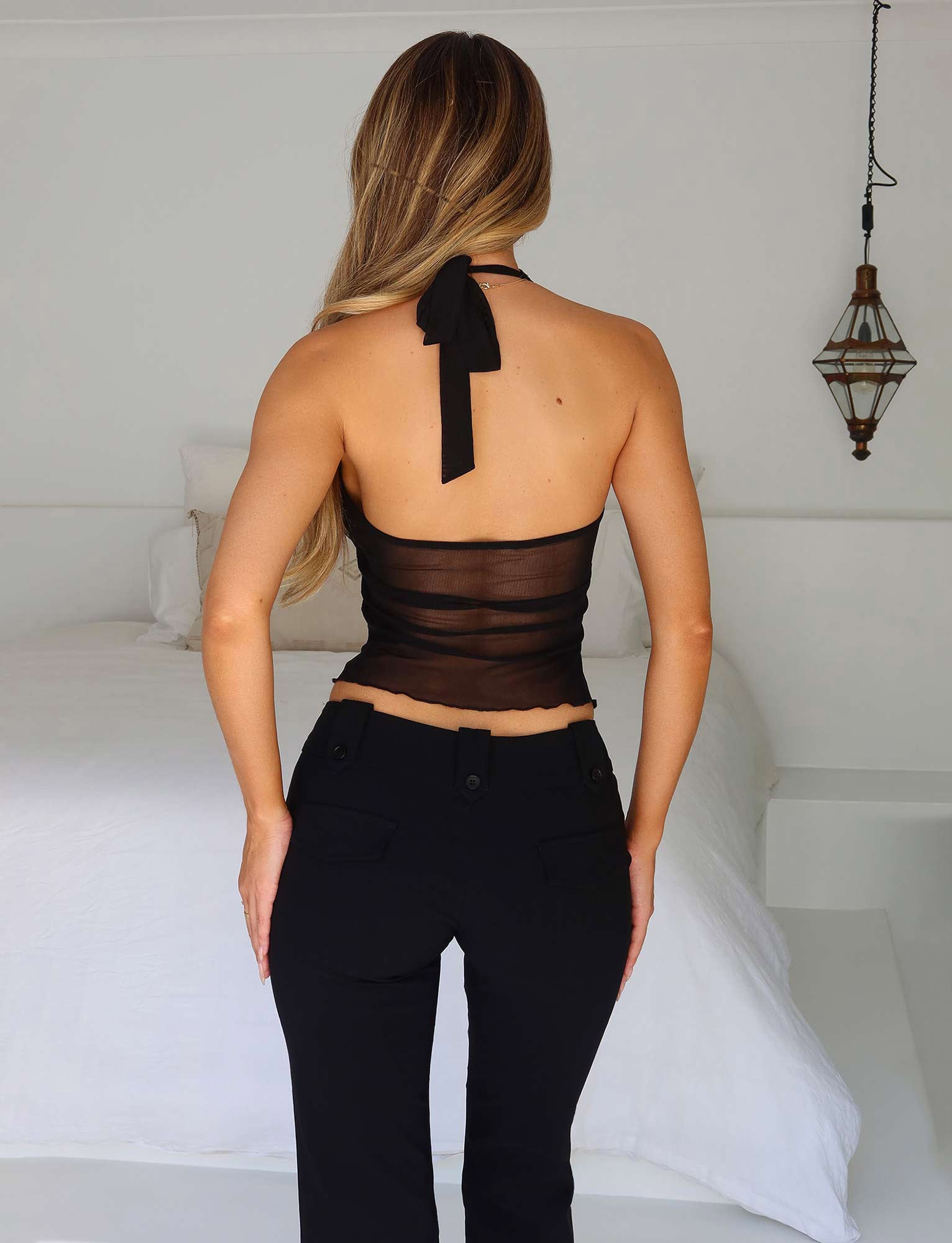 A woman with long, blonde hair is standing with her back to the camera in a minimalistic white bedroom. She is wearing the KAYLEE TOP - BLACK by TIGERMIST, featuring a mesh back, paired with black high-waisted pants. A single hanging light fixture is visible on the right side of the image.