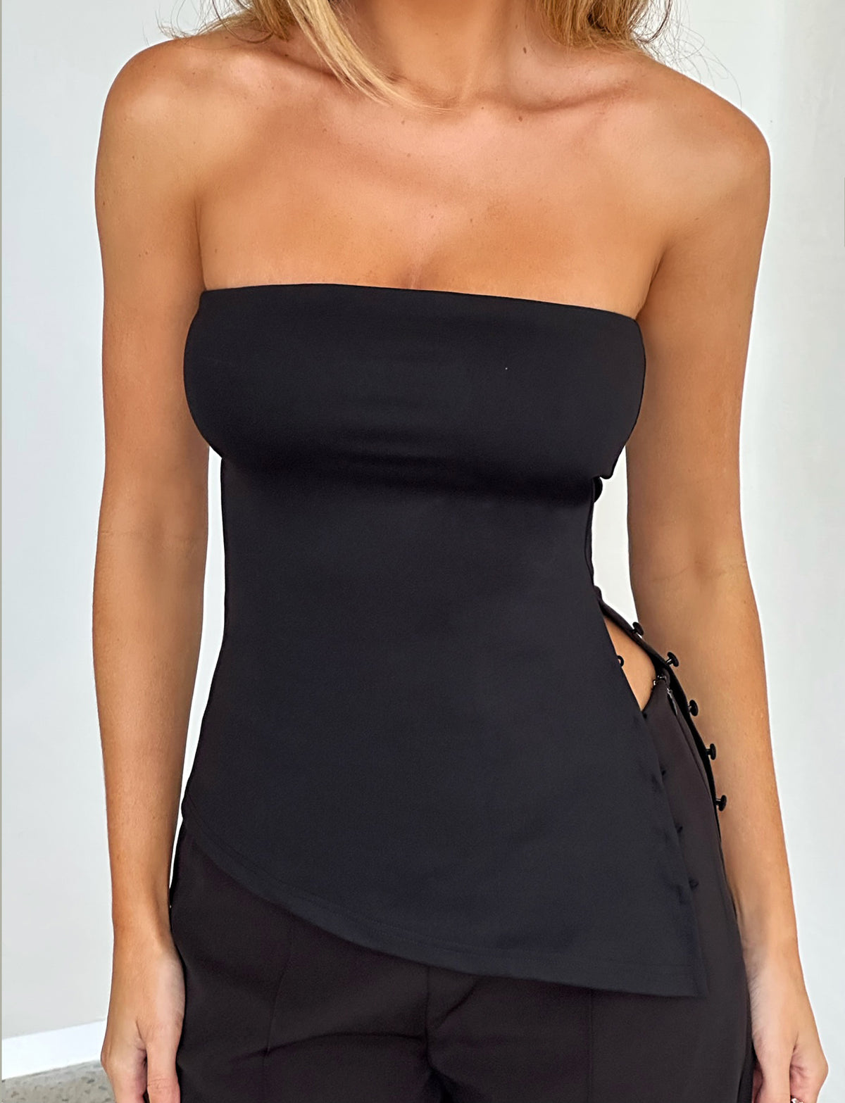 A person is wearing the INDIRA TOP - BLACK from TIGERMIST, featuring a strapless design with an asymmetrical hemline and side-button details. The top is paired with black pants against a plain white background.