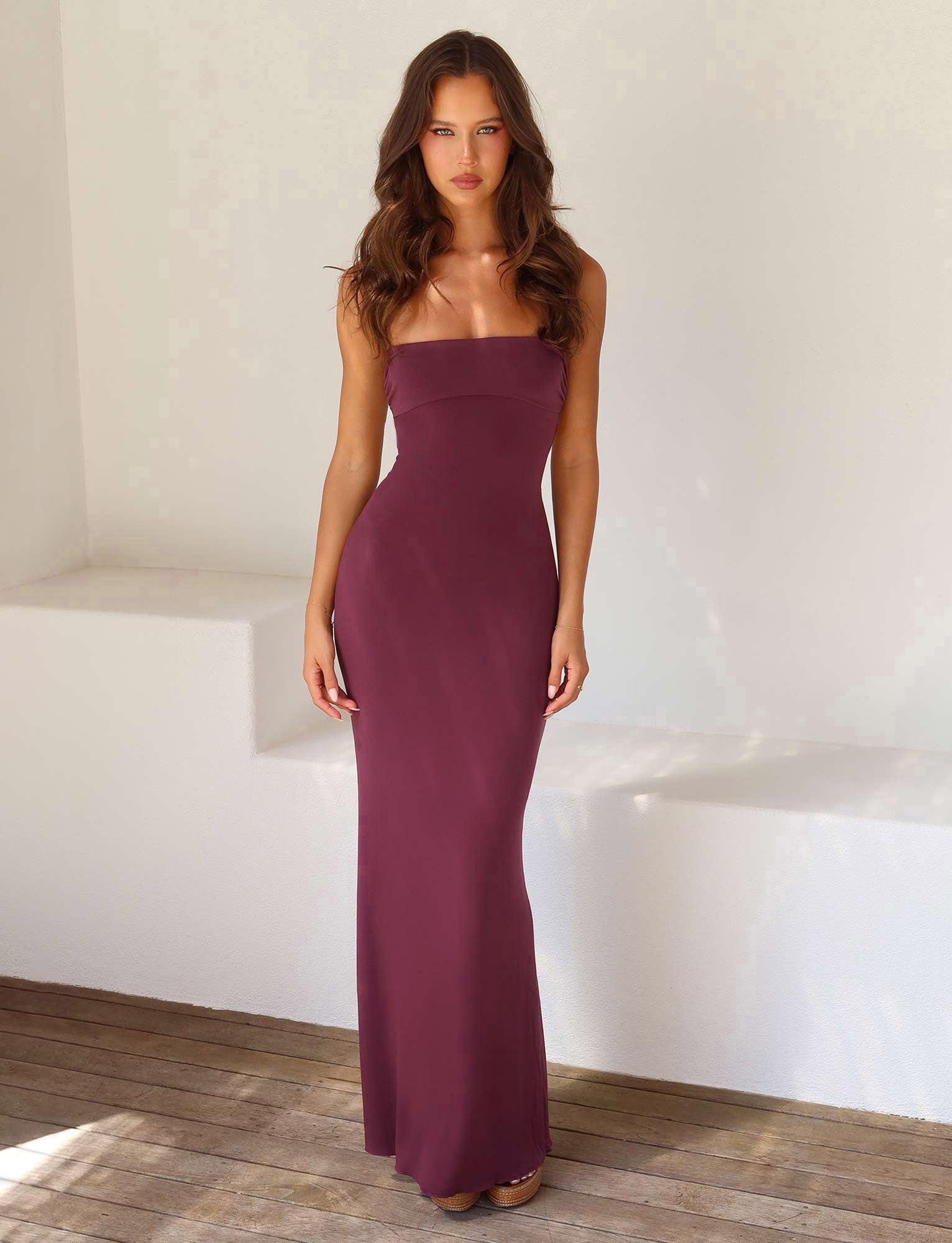 A woman with long, wavy hair stands against a plain white background, wearing the form-fitting, strapless CARA DRESS in Raisin from TIGERMIST that reaches the floor. She is looking directly at the camera with a neutral expression and is posed on wooden flooring.