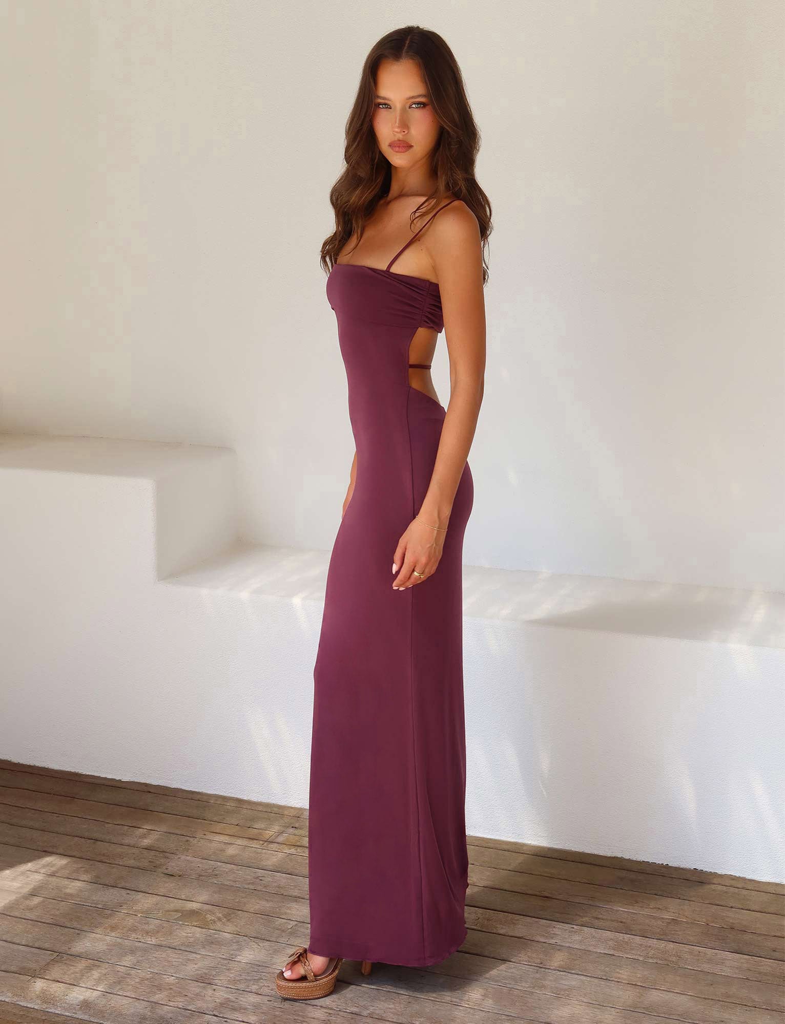 A woman stands on a wooden floor against a white wall. She has long wavy hair and wears the form-fitting, sleeveless CARA DRESS - PURPLE : RAISIN from TIGERMIST, featuring thin straps and side cut-outs. She is looking directly at the camera, with a few strands of hair falling over her shoulders.