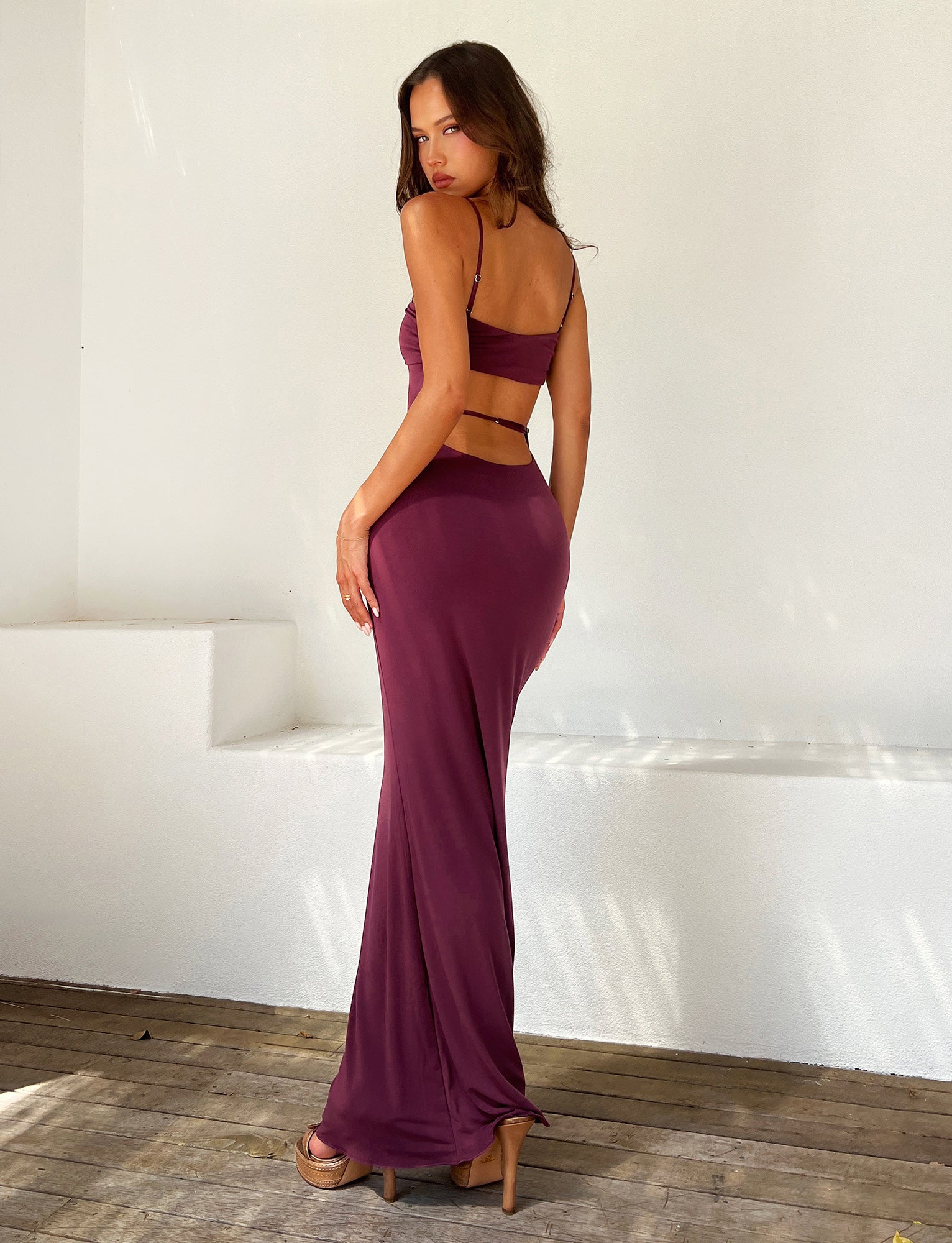A woman stands on a wooden floor against a white wall, facing sideways and looking over her shoulder. She is wearing the form-fitting, backless CARA DRESS - PURPLE : RAISIN from TIGERMIST with spaghetti straps and high heels. Light casts soft shadows around her.
