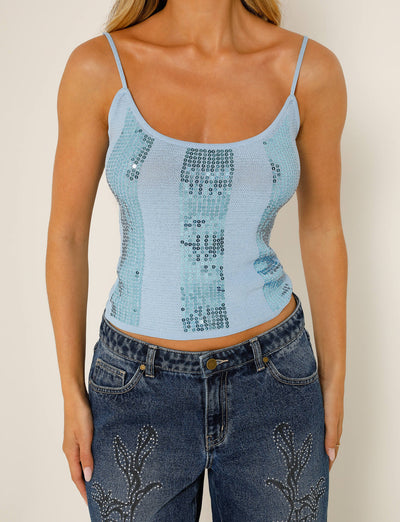 A person wearing the JUNNO TOP - BLUE from TIGERMIST, paired with blue jeans featuring embroidered detailing on the thighs, stands against a neutral background.